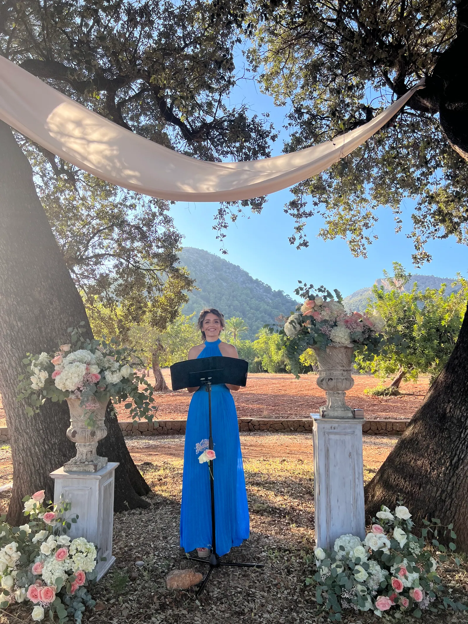 Ximena standing in an outdoor ceremony setting beneath large trees and white drapery