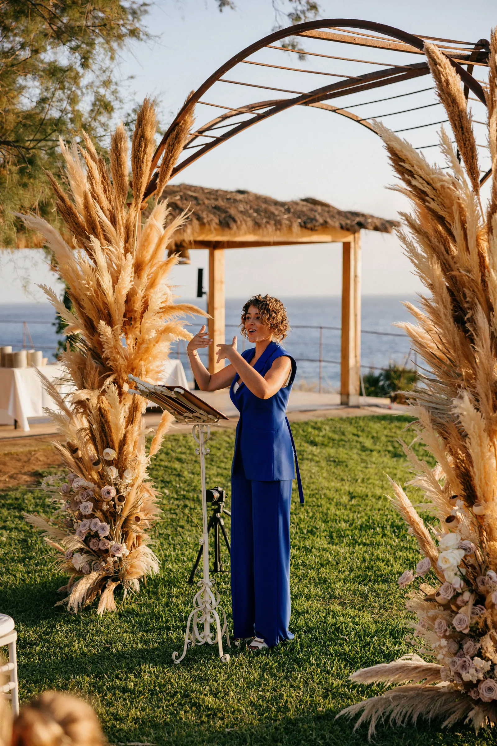 Ximena speaking expressively beneath a sculptural ceremony arch beside the water