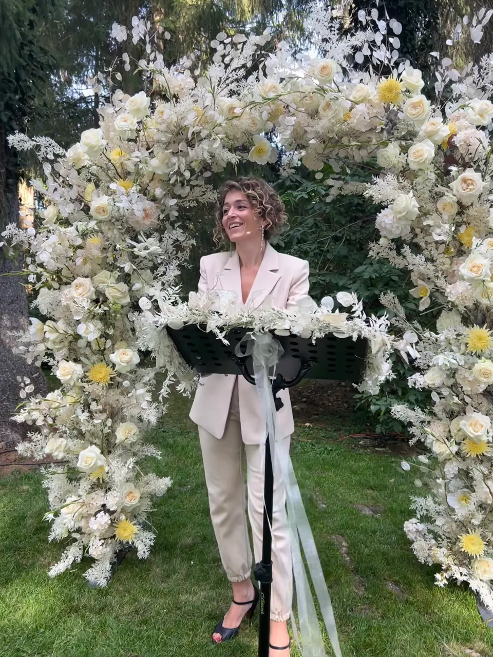 Ximena smiling at a lectern beneath an arch of white flowers