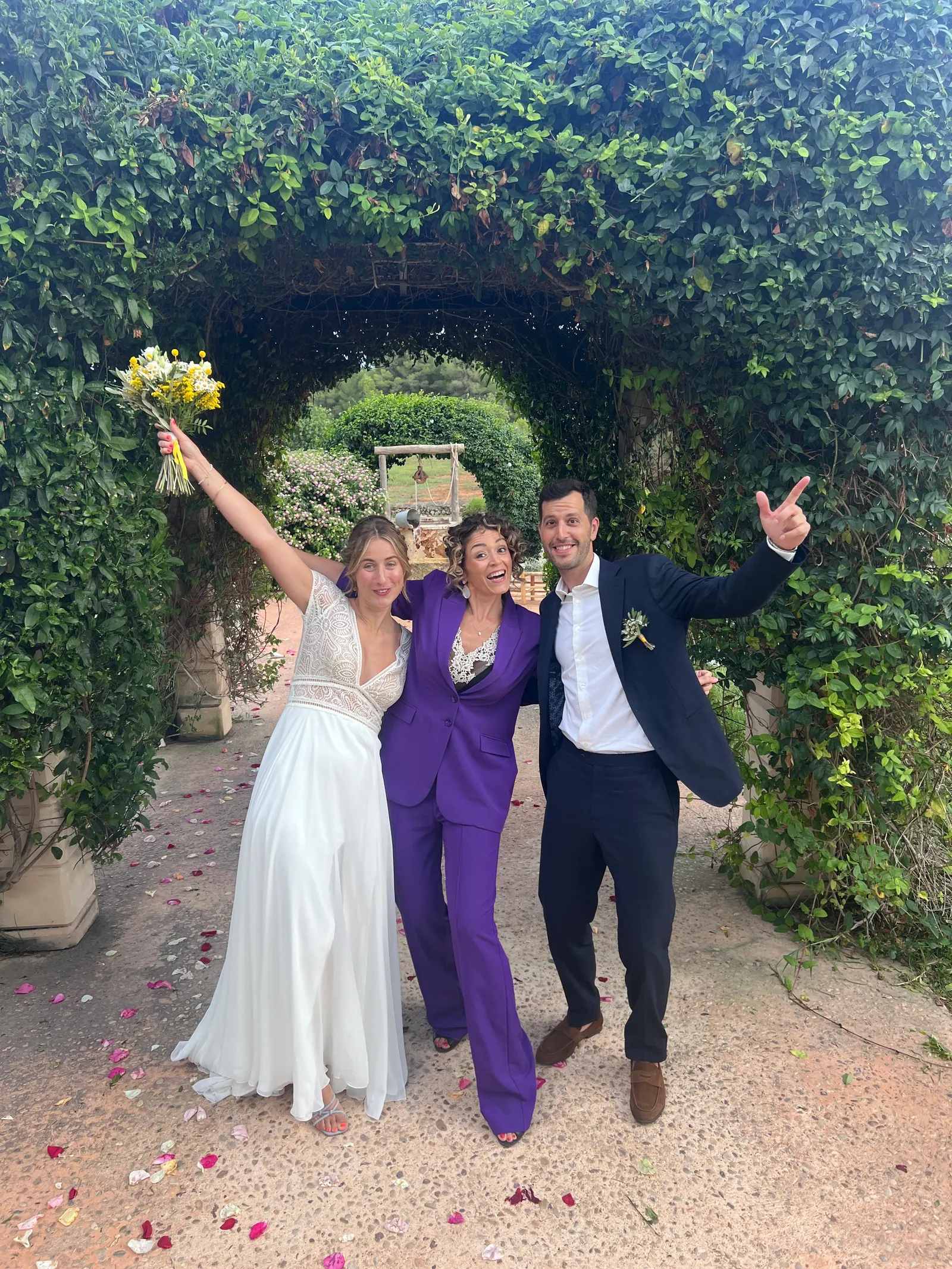 A couple and Ximena raising their arms beneath a green garden arch