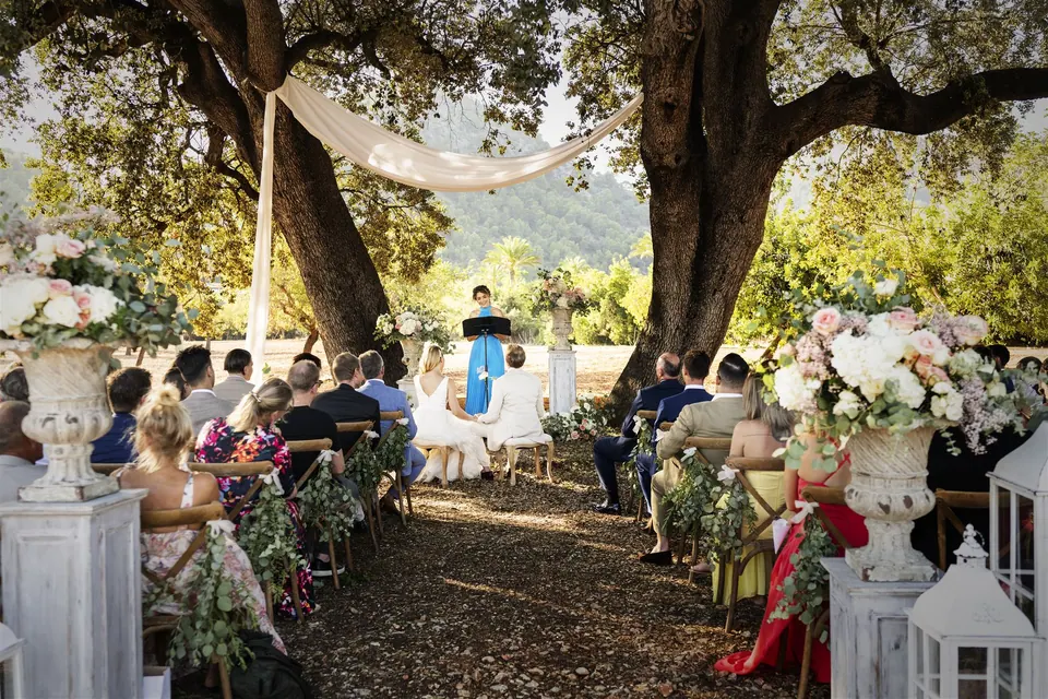 A couple holding hands during a ceremony led by Ximena, surrounded by their guests under large trees