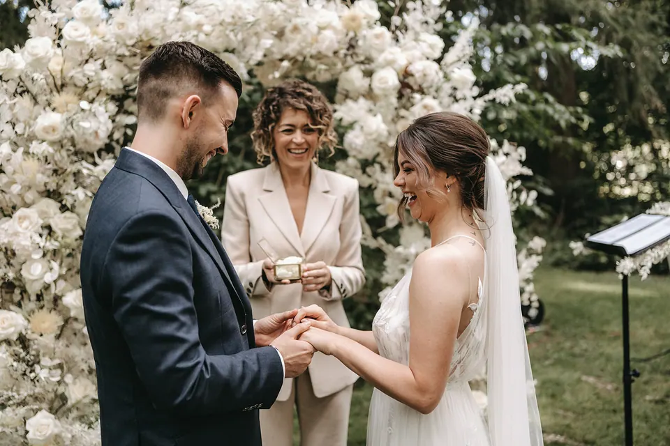 A couple exchanging rings while Ximena laughs with them beneath white flowers