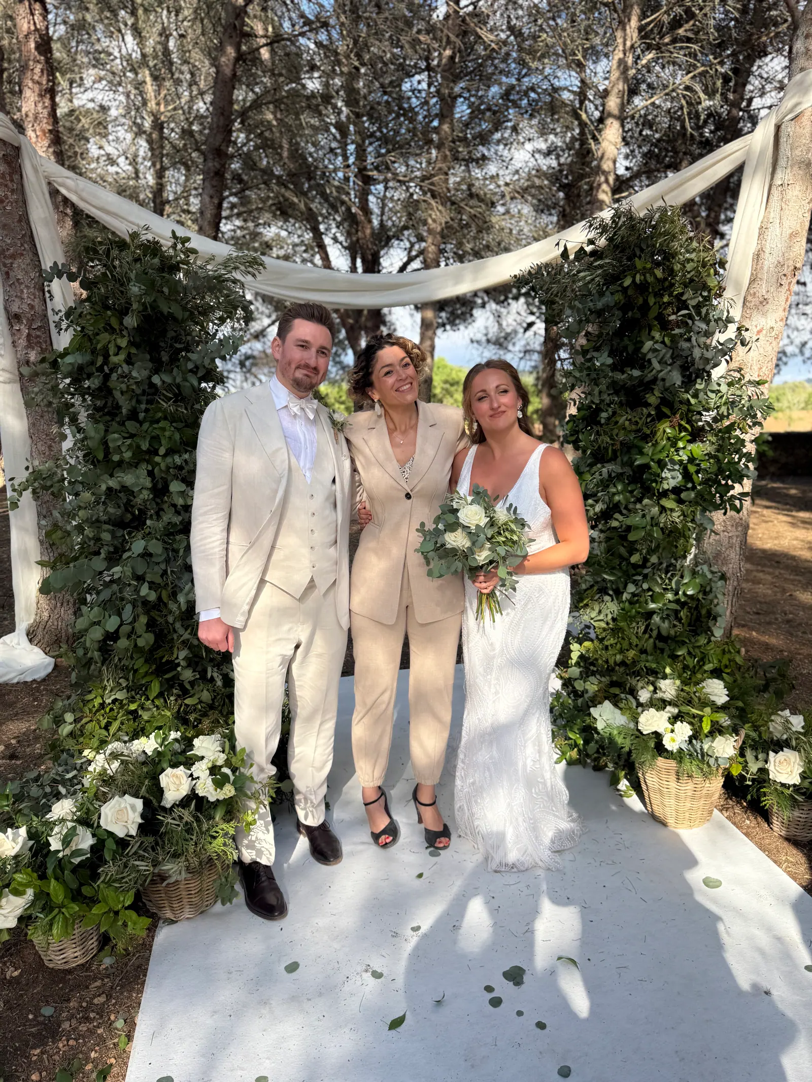 A couple standing with Ximena beneath trees and white flowers after their ceremony