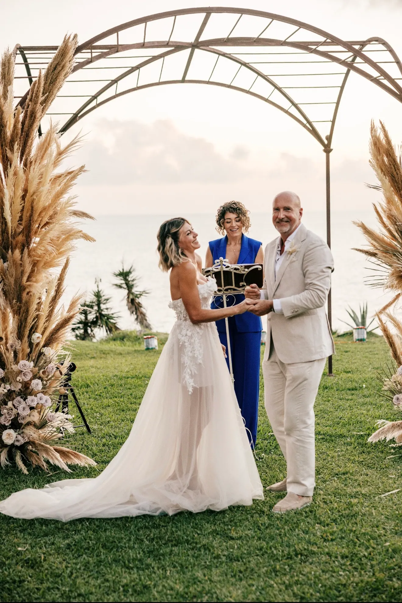 Ximena leading a wedding ceremony beneath a sculptural arch beside the water