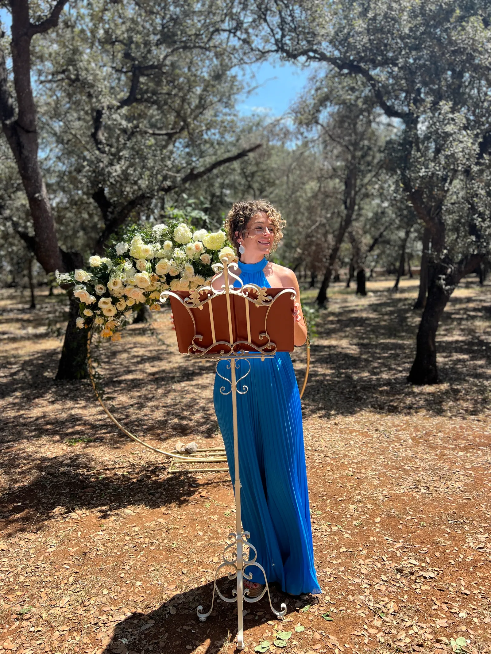 Ximena smiling with her ceremony book beneath trees and white flowers