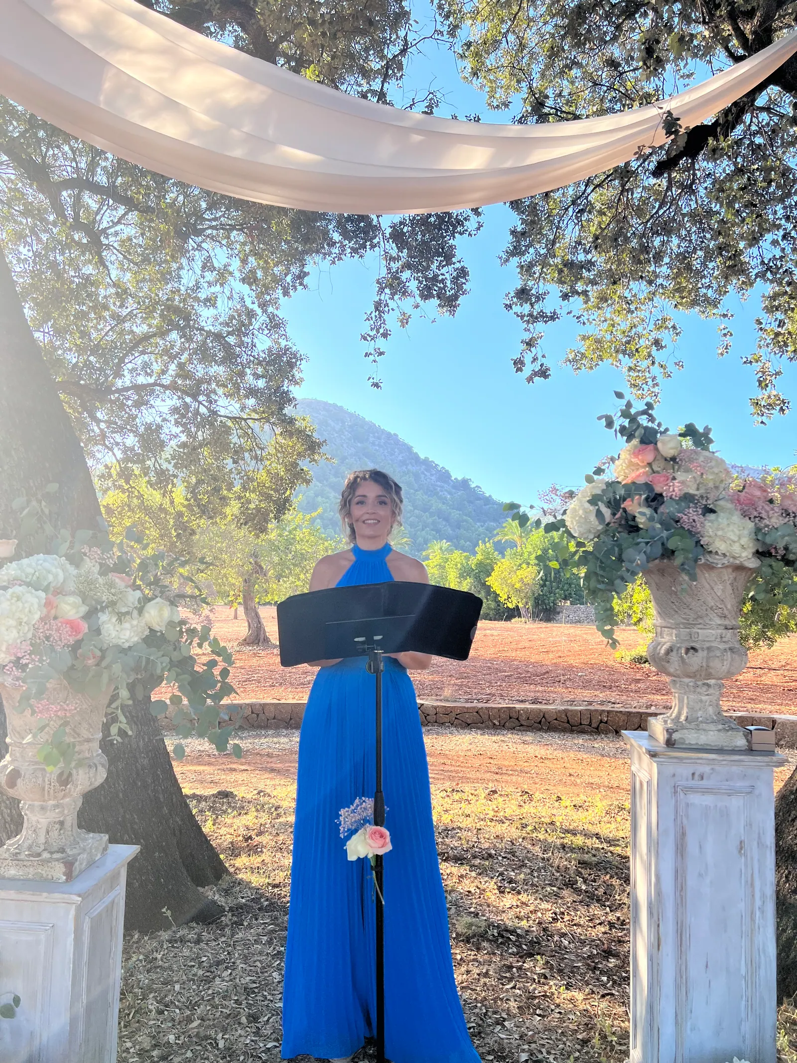 Ximena smiling at a lectern beneath trees, flowers and white drapery
